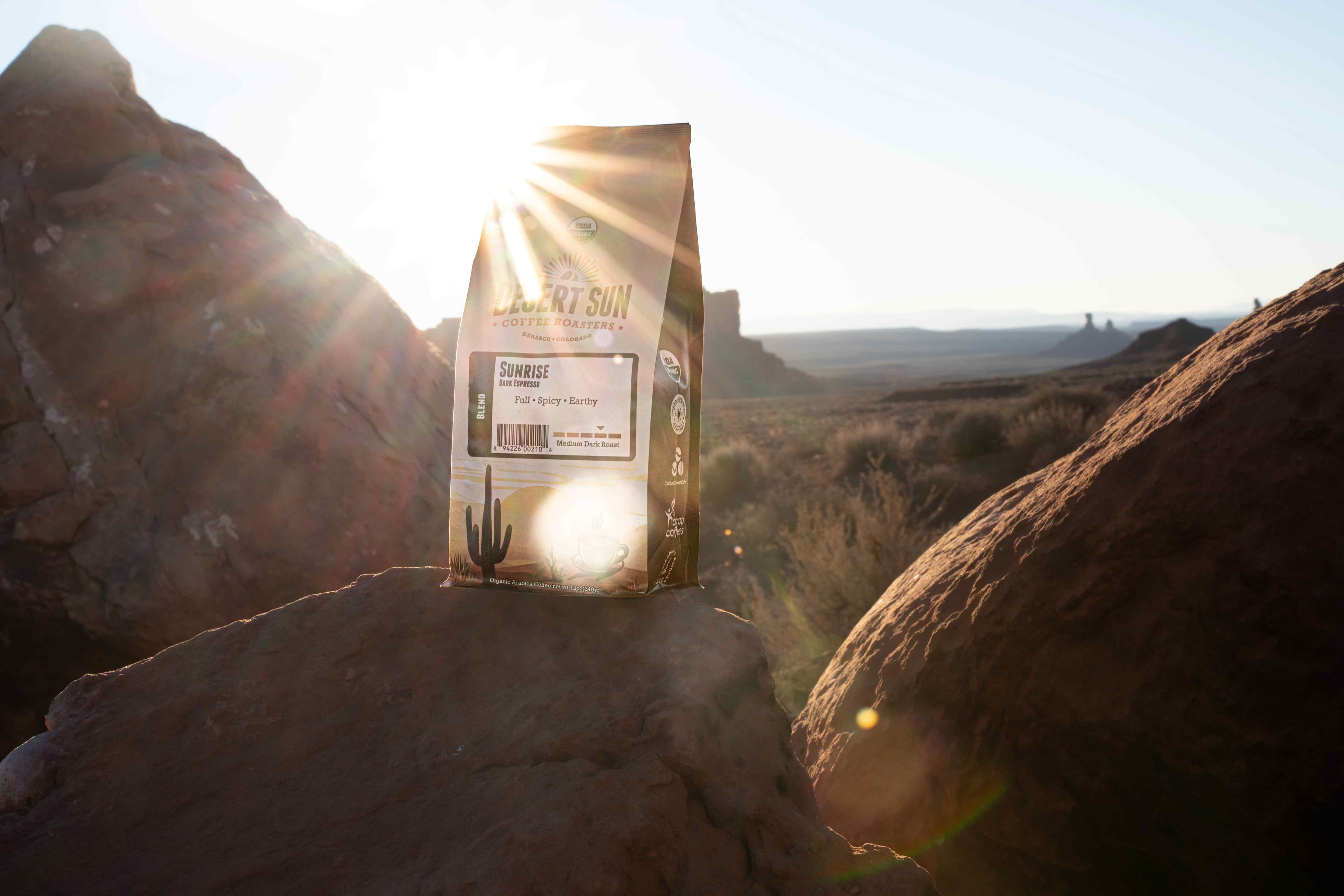 A bag of Desert Sun coffee between boulders with the sun bursting over its shoulder