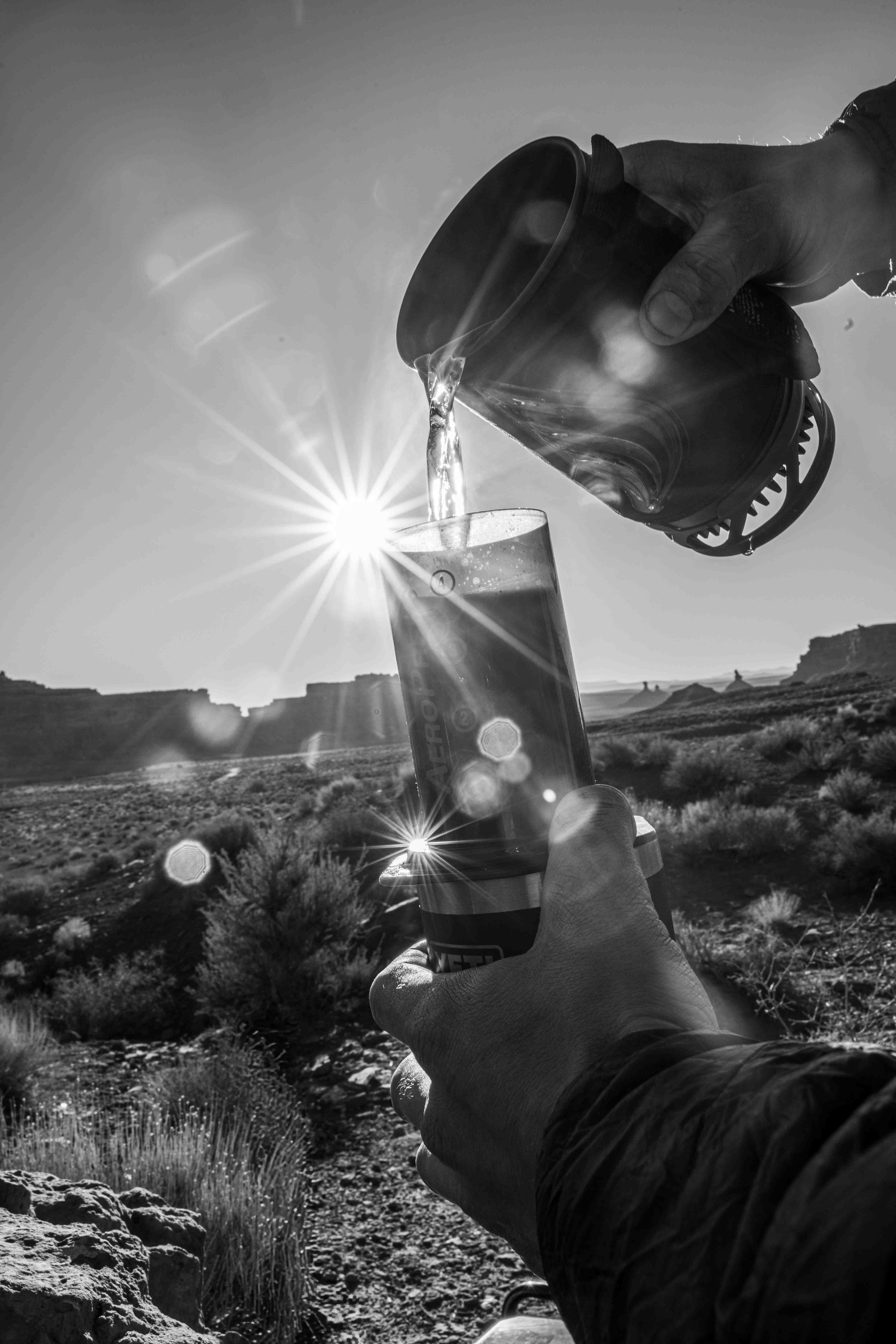 Black-and-white photo of coffee pouring from a kettle, the rising sun flaring behind it over the desert
