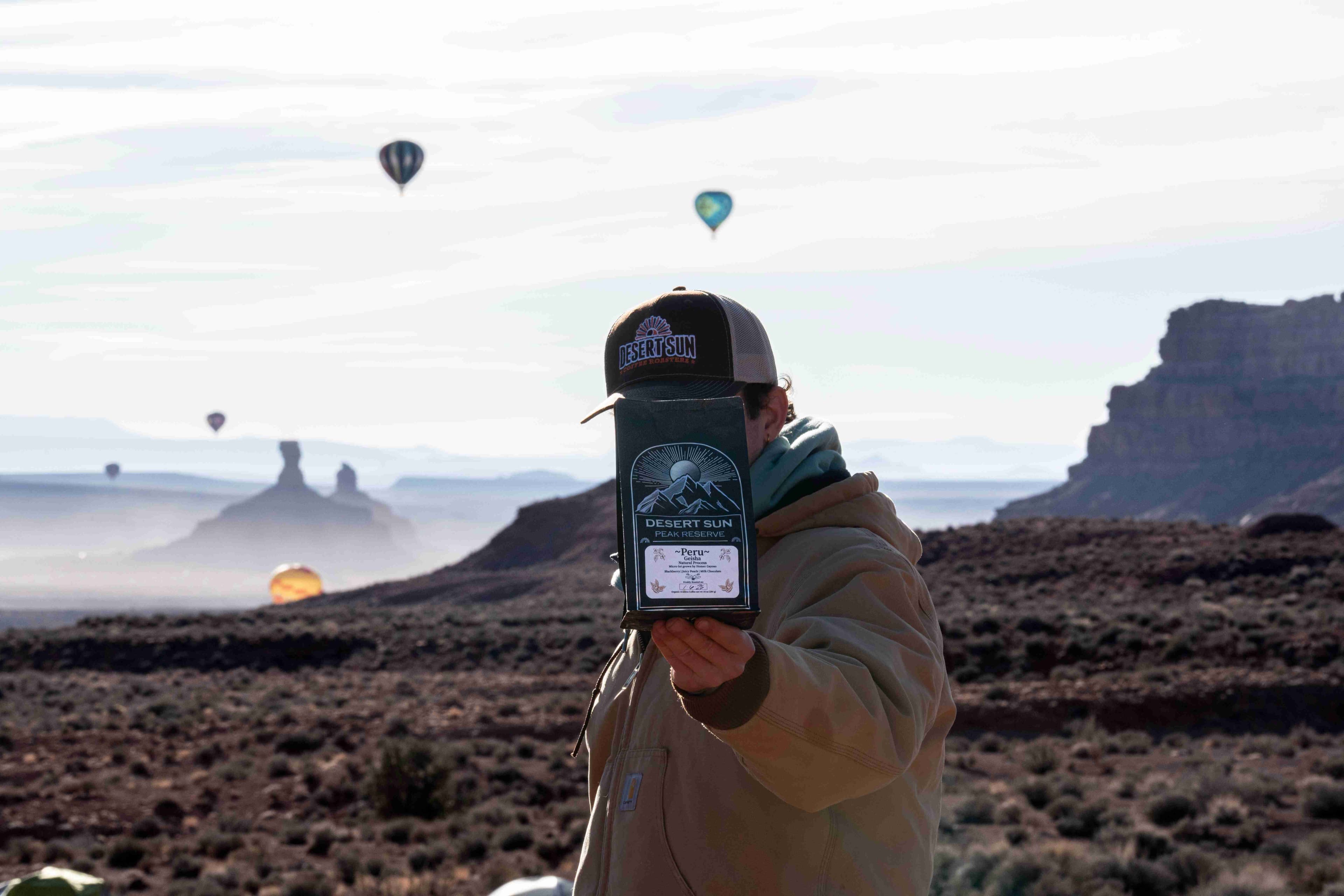 A man holds a bag of Desert Sun Peak Reserve up against the sky, balloons drifting over the buttes behind him