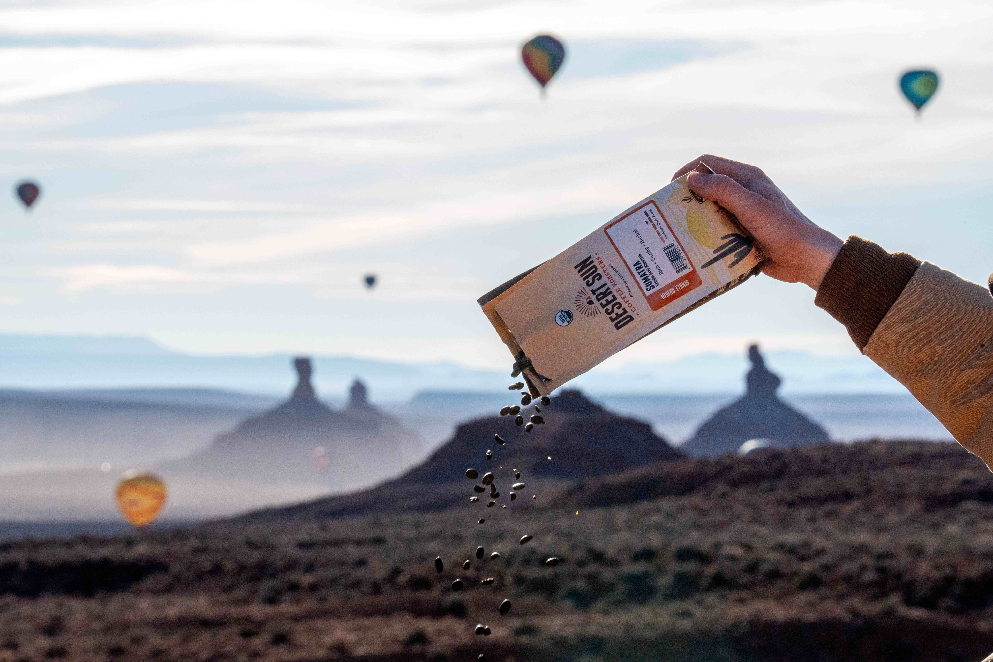 Coffee beans pouring from a bag of Desert Sun Sumatra over the Valley of the Gods as hot air balloons drift above the buttes