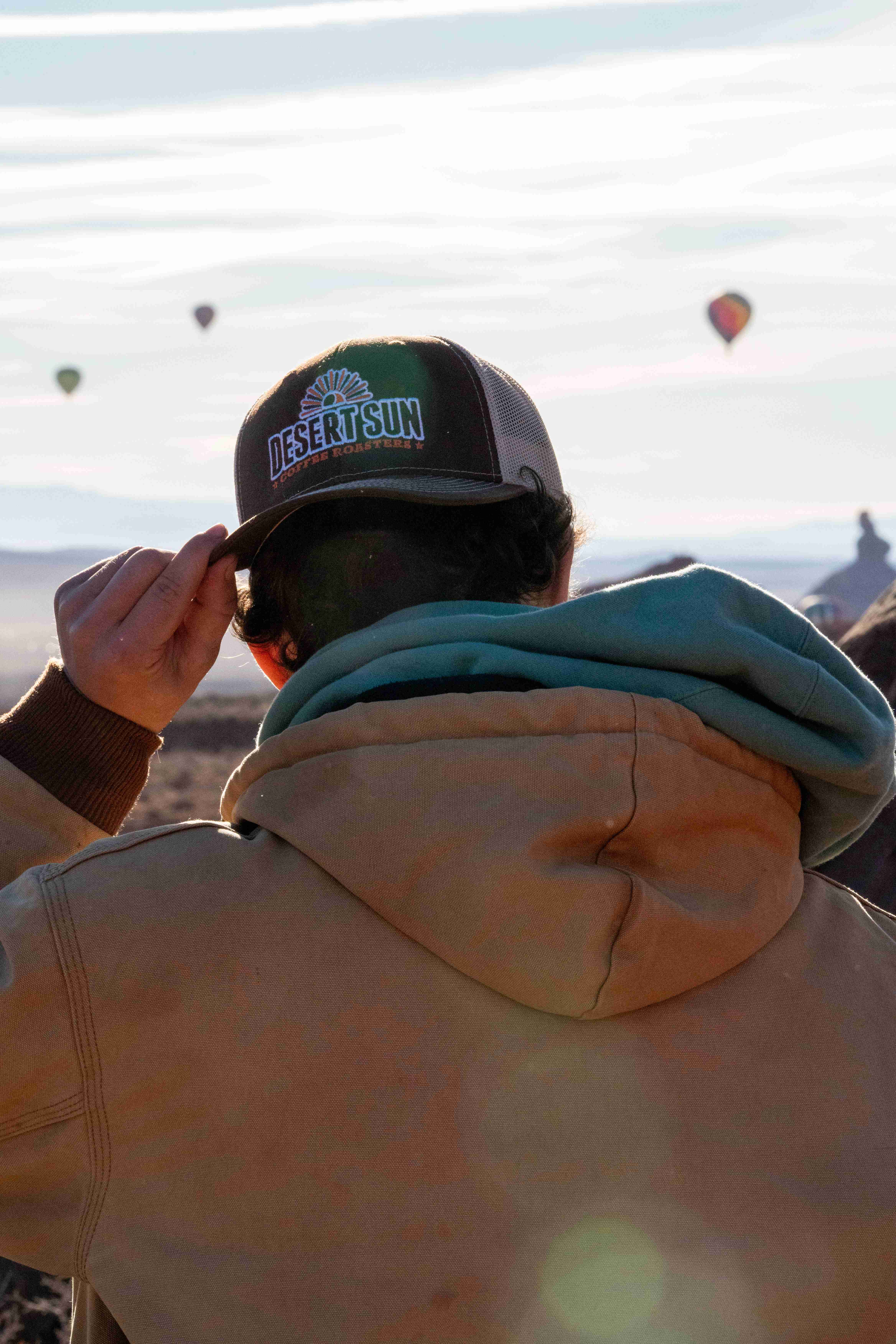 A man seen from behind adjusting his Desert Sun cap as balloons float over the valley
