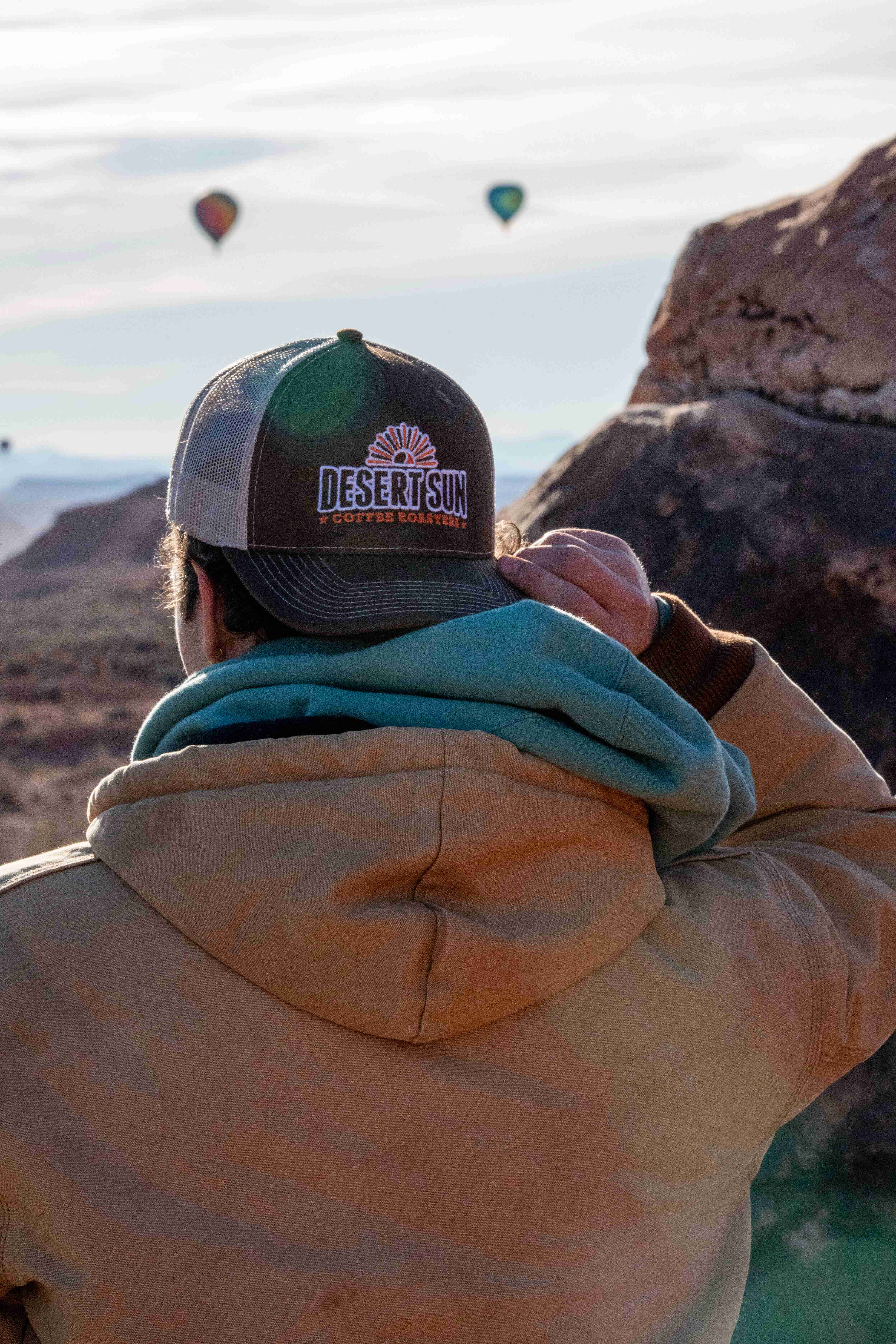A man in a Desert Sun cap and hooded jacket drinks from a mug, looking out at the balloons