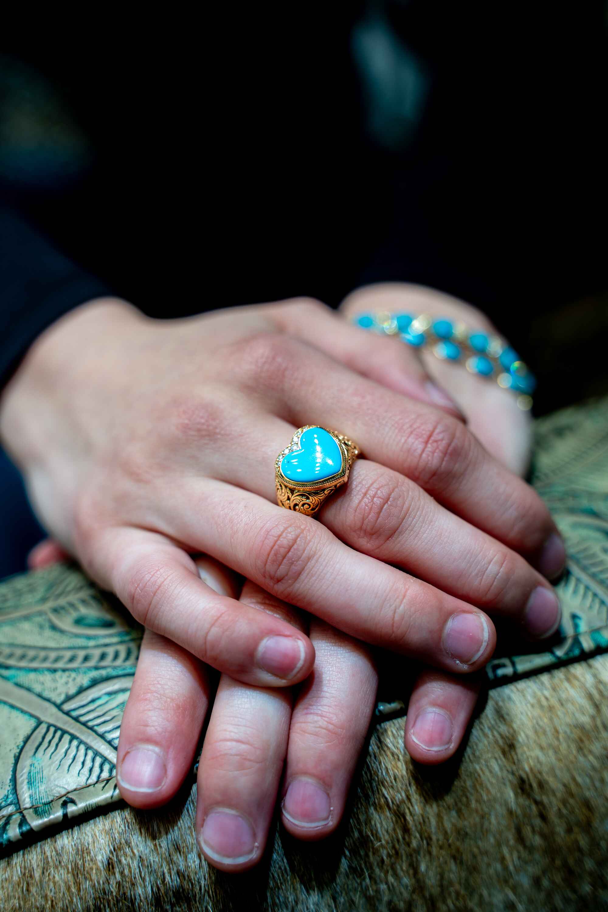 A turquoise heart ring and turquoise bracelet from Feathers Galleria on the Plaza, hands resting on tooled leather and hide — a Creative Flow Studio frame from Santa Fe.