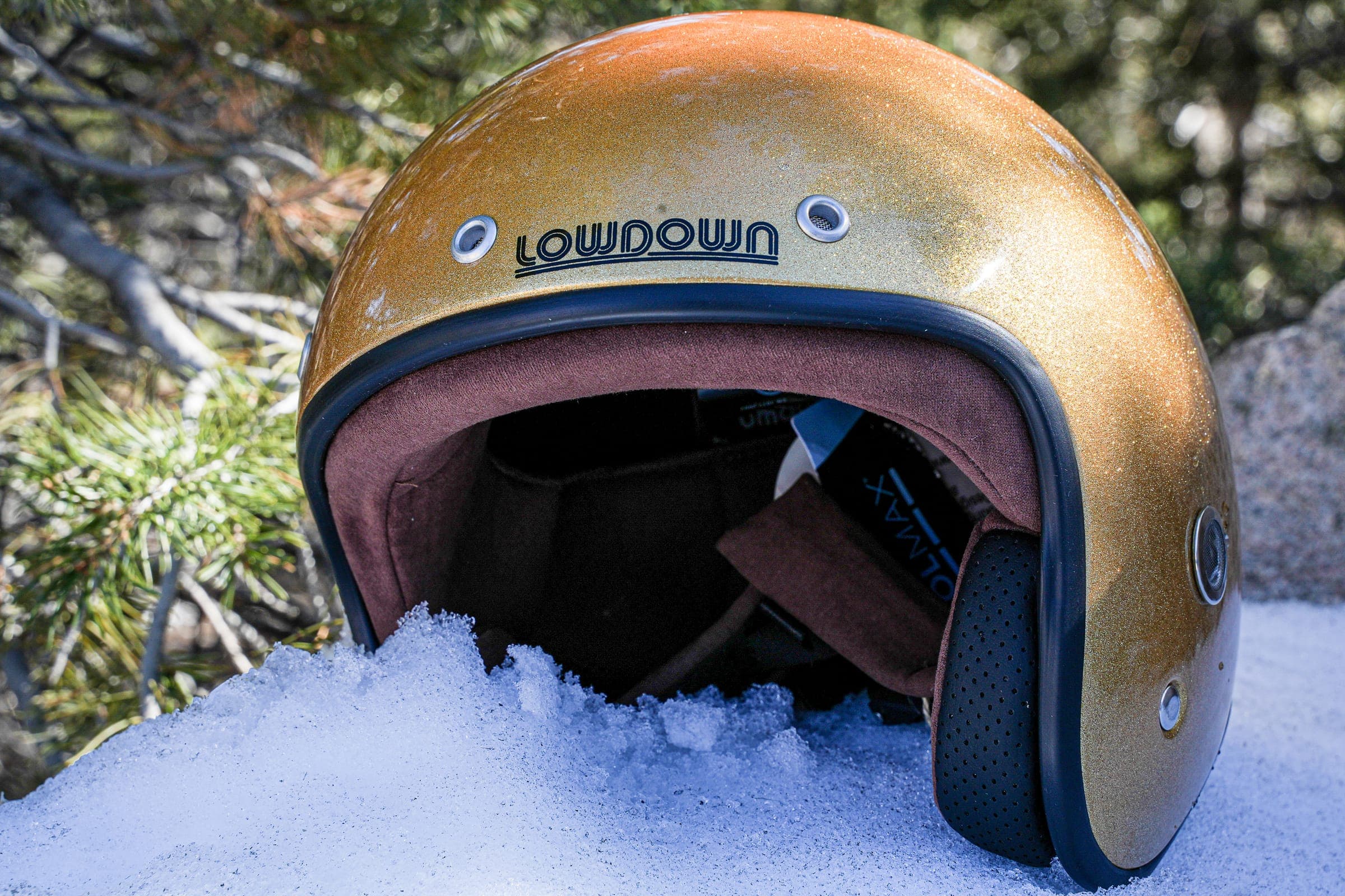 A gold glitter Lowdown helmet set into fresh snow at the base of a pine, its brown leather lining showing