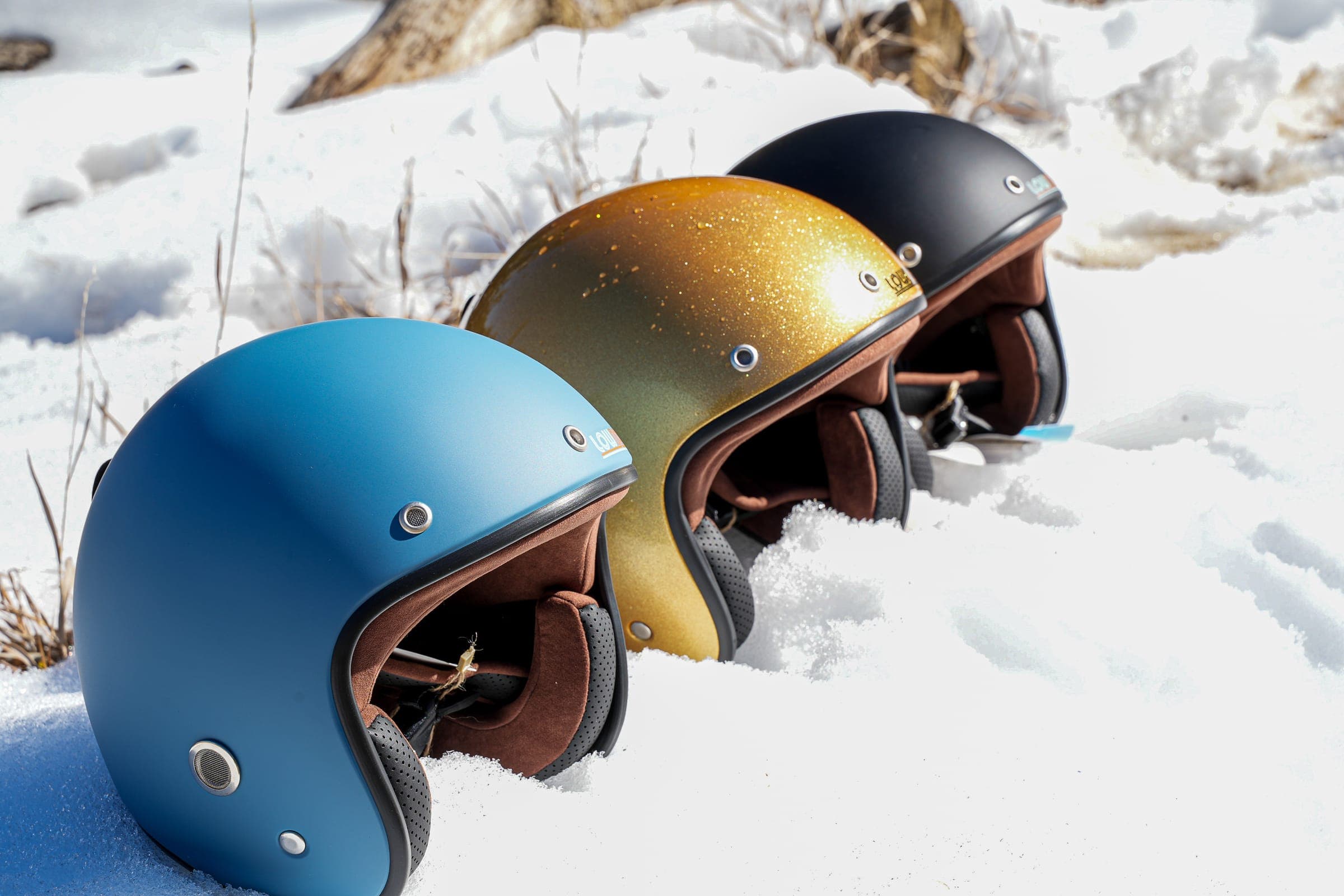 Three Lowdown helmets, blue, gold glitter, and black, set into fresh snow