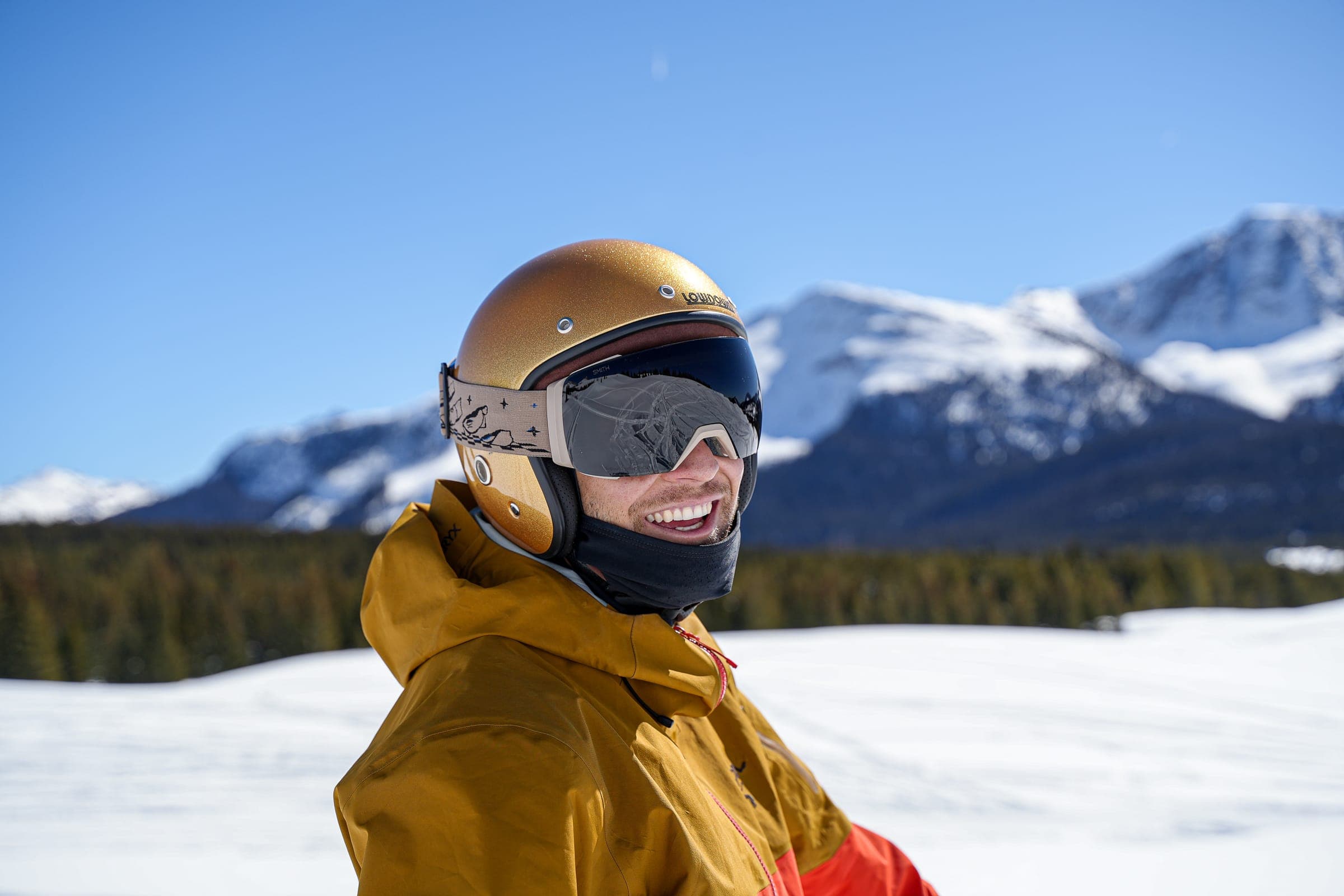 A skier in a gold helmet and goggles looking off to the side, a range of white peaks behind