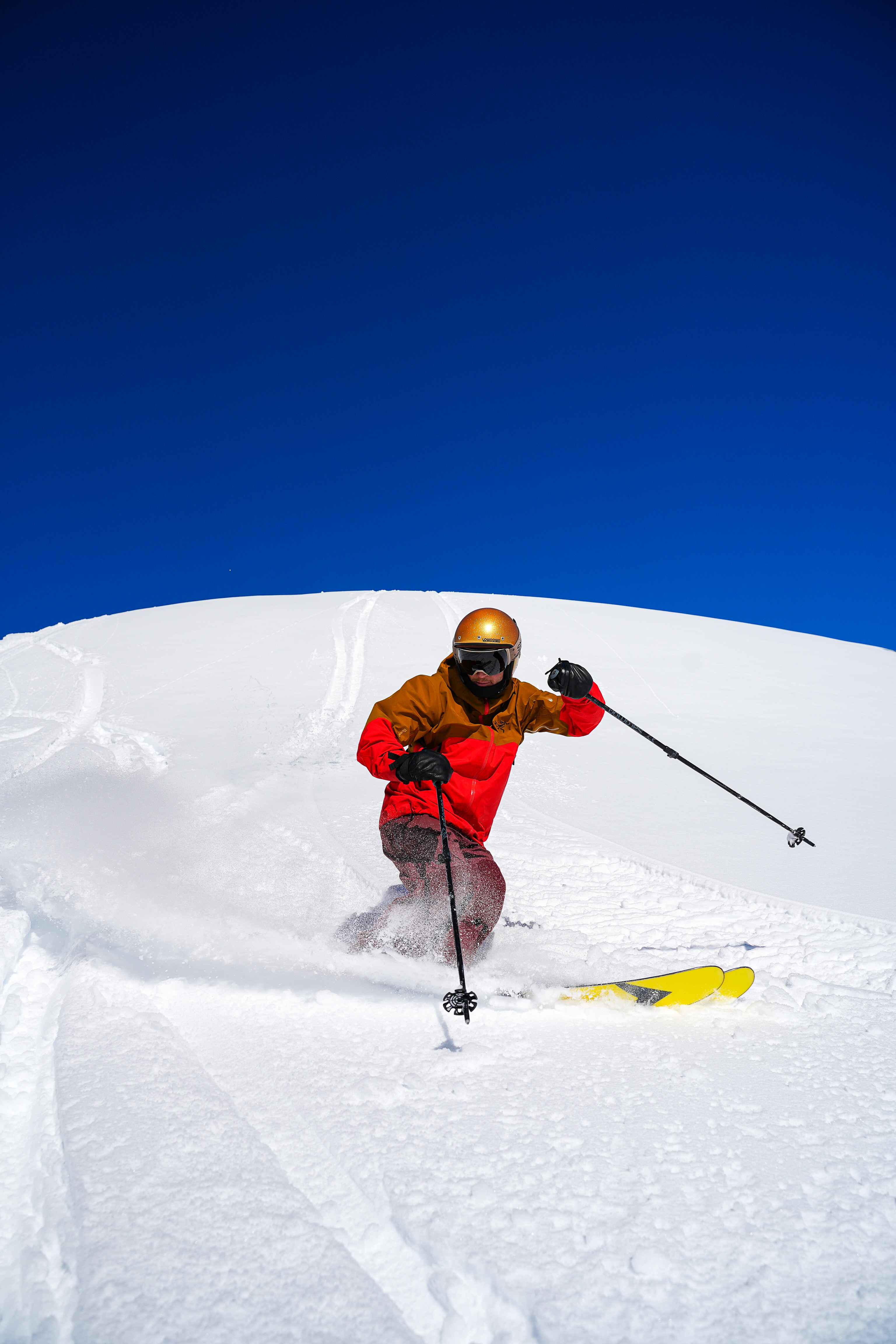 A skier in a gold helmet and red jacket turning through untracked snow under a deep blue sky