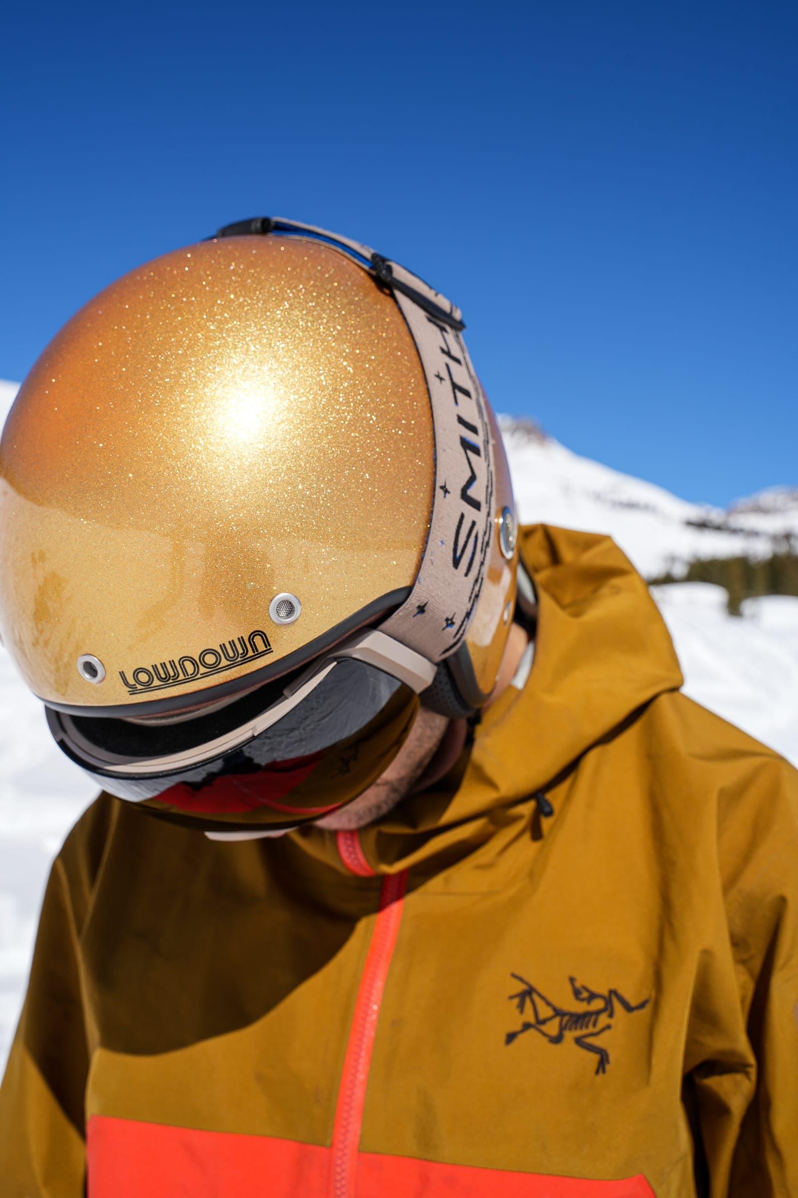 Close-up of a gold glitter Lowdown helmet and goggles on a skier in an orange jacket