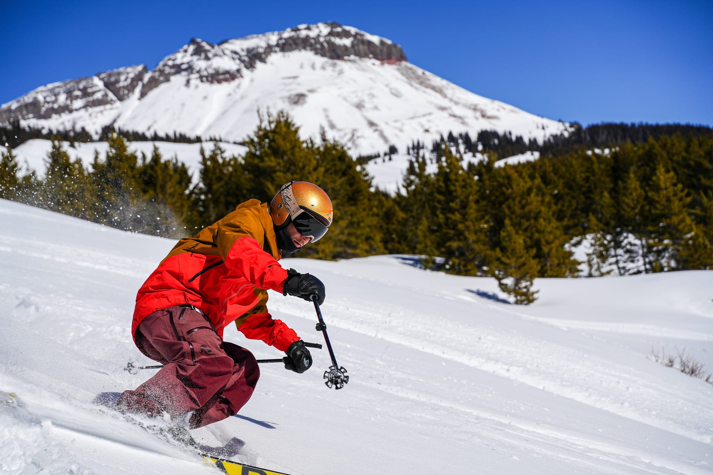 A skier in a red jacket and gold helmet turning hard in powder, a snow-capped peak and pines behind