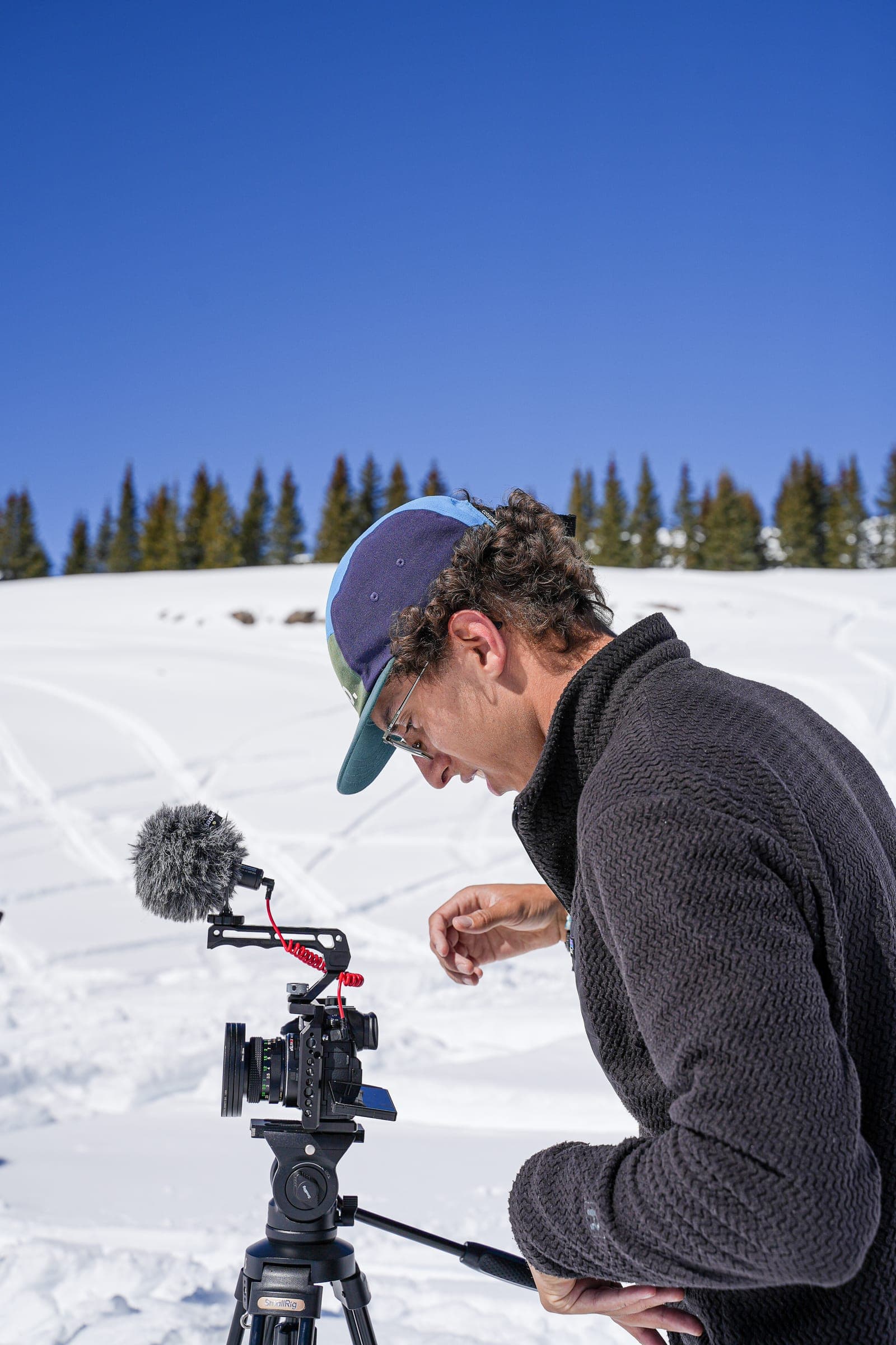A crew member in a black jacket setting up a video camera with a shotgun mic on a tripod in the snow
