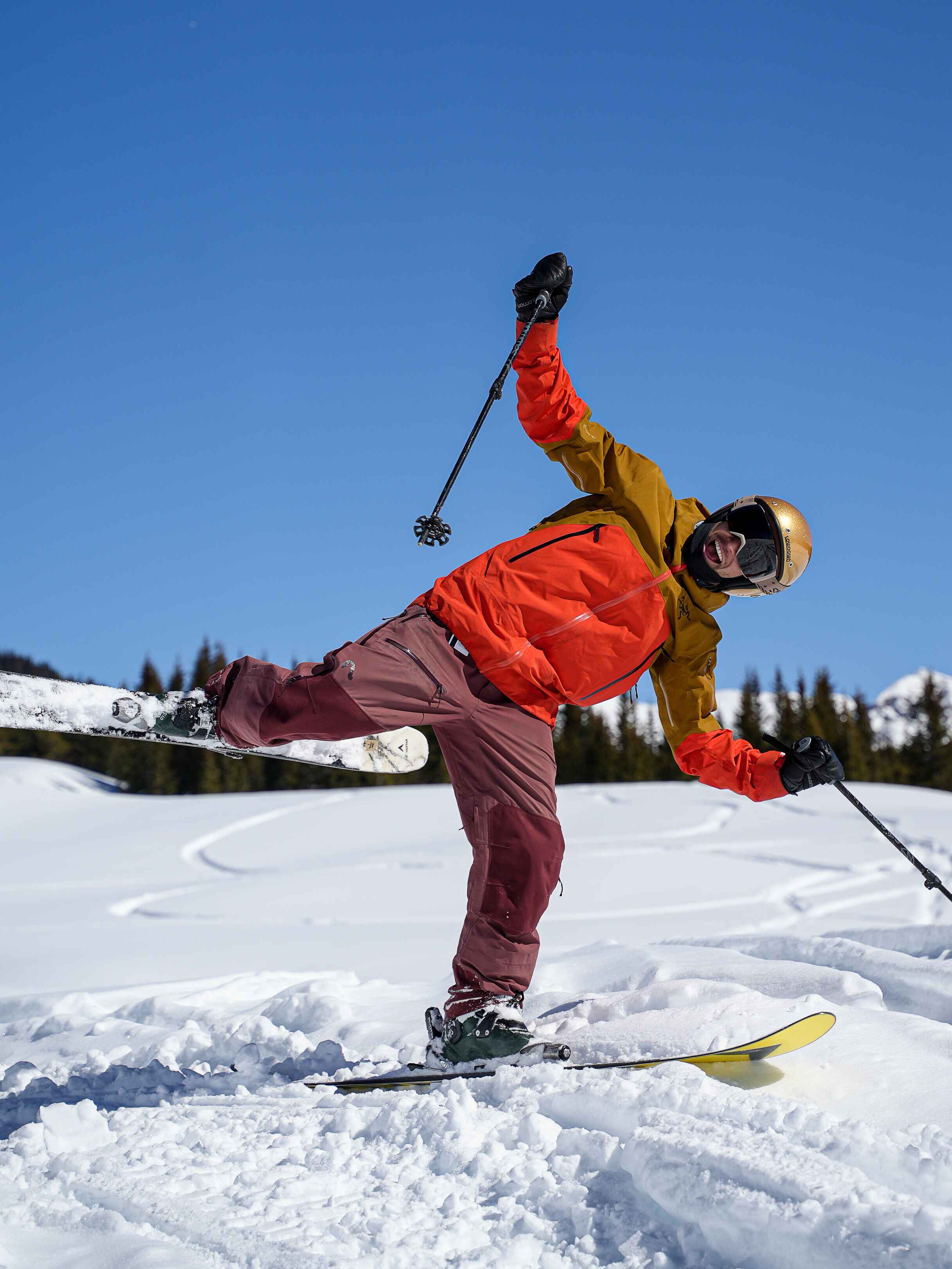 A skier in a gold helmet striking a playful one-ski pose with a pole raised, mountains behind