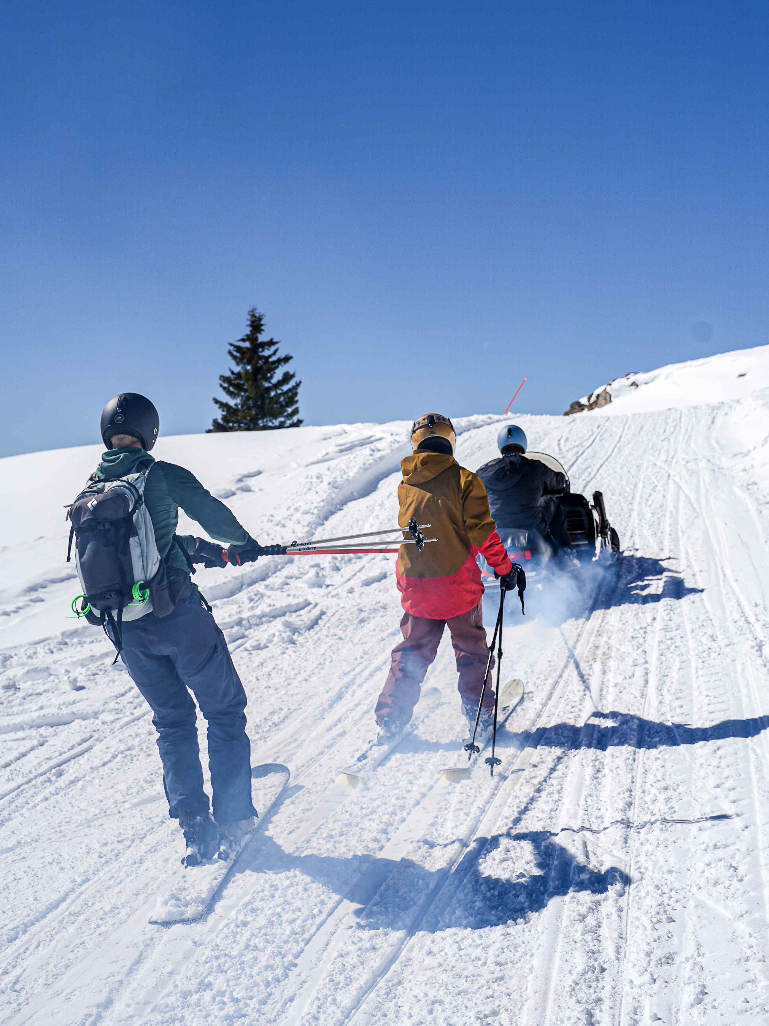 Two skiers being towed across a snowy track, one in a gold helmet, kicking up snow