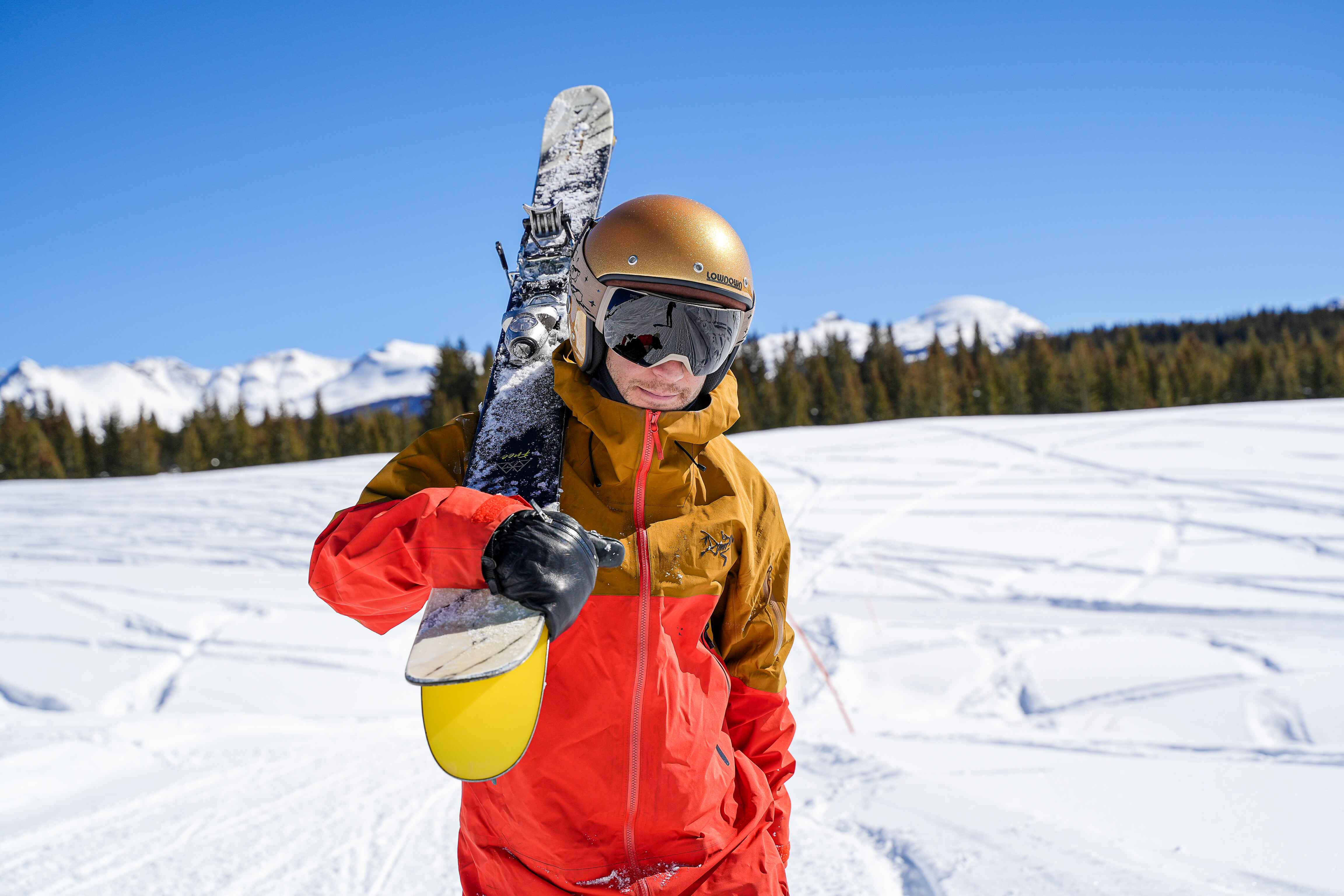 A skier carrying skis in a gold glitter Lowdown helmet with snowy peaks behind — brand storytelling engagement