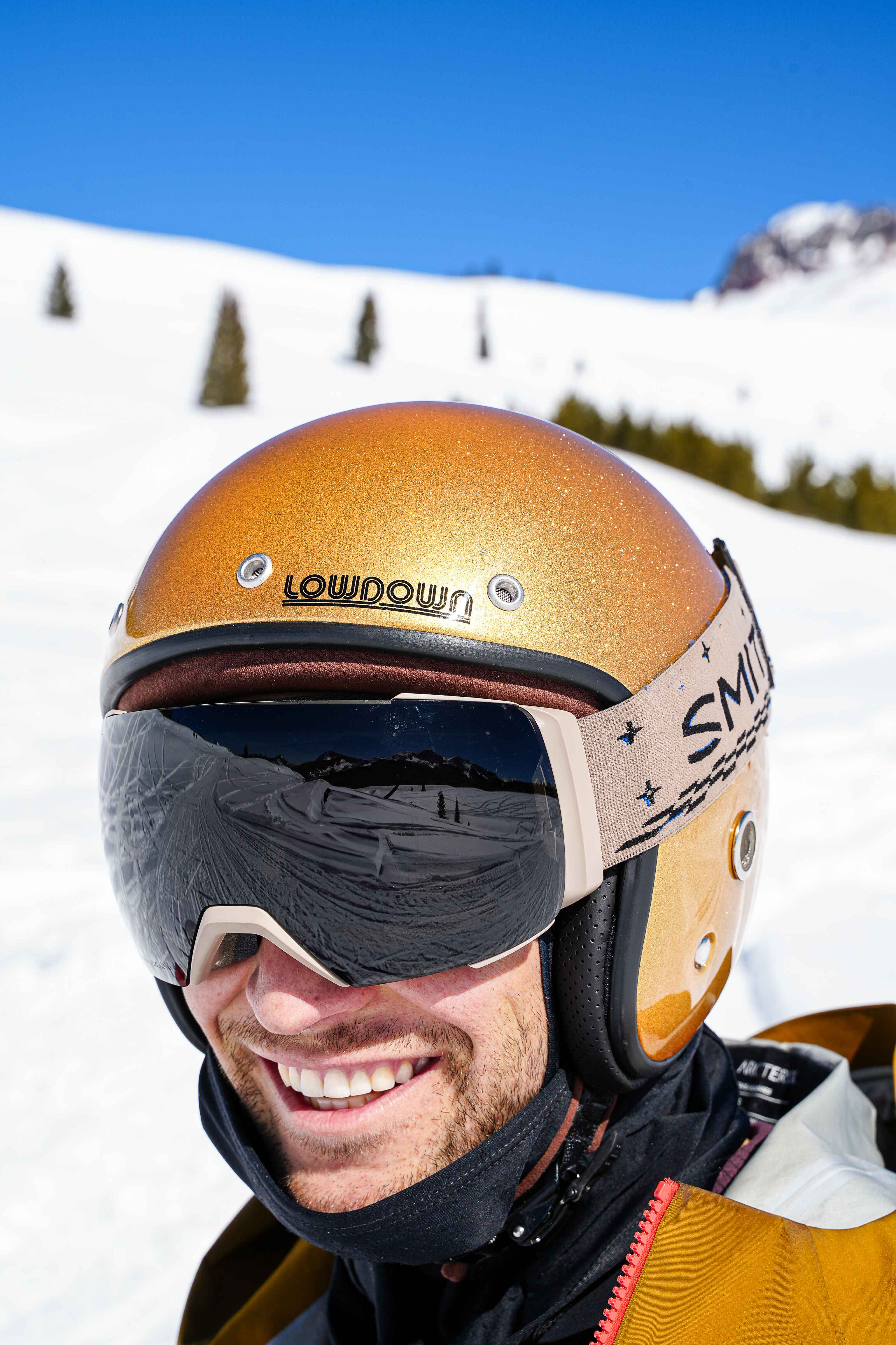 A grinning skier in a gold Lowdown helmet and goggles, close up, snow and blue sky behind