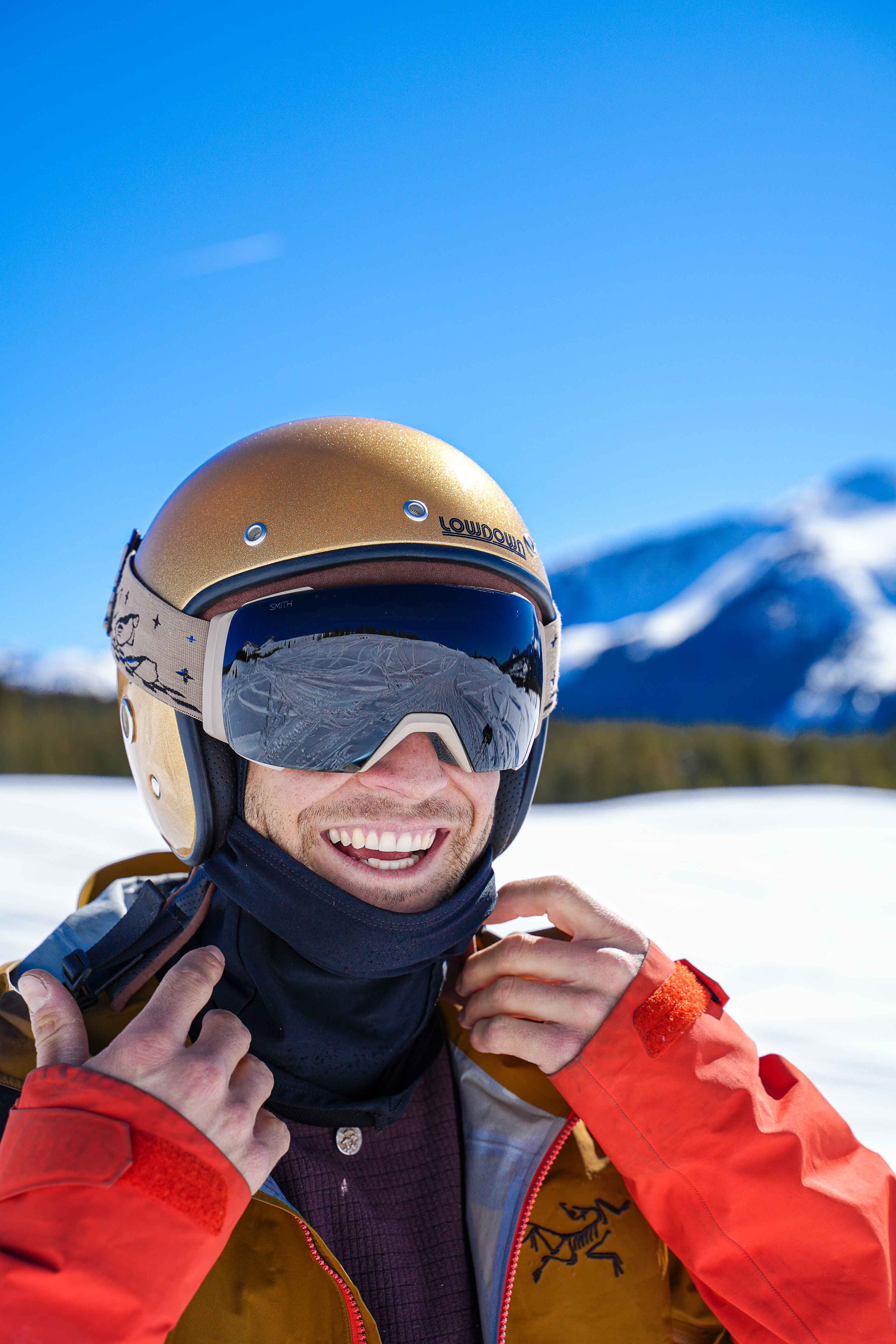 A smiling skier in a gold helmet and goggles pulling up the collar of a red jacket, mountains behind