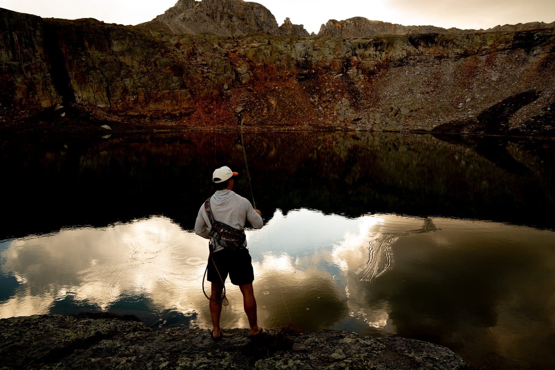 An angler standing on the rocks of a still lake that mirrors the cliffs and sunset clouds
