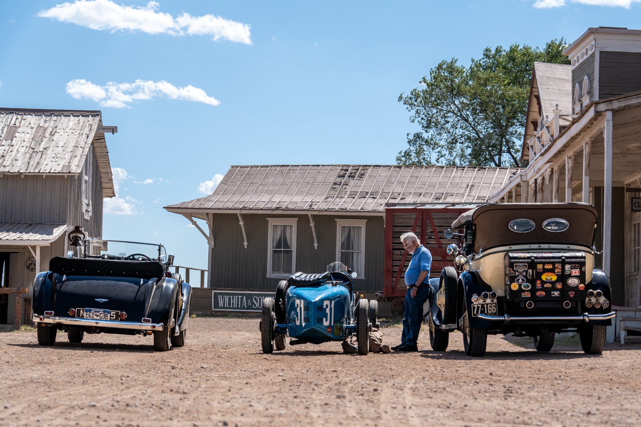 Three vintage cars parked on the dirt main street of a western movie town near Santa Fe while their owner checks a blue Bugatti — a Creative Flow Studio frame from Eaves Movie Ranch.