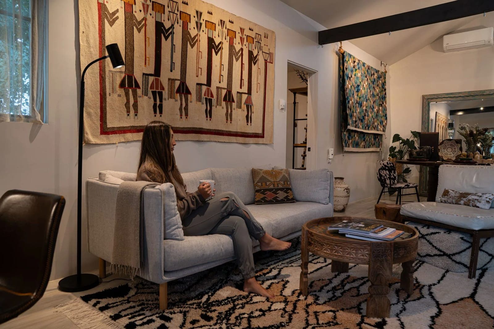 A guest with a mug on the sofa in the Farmhouse 2 living room, under a woven textile of dancing figures
