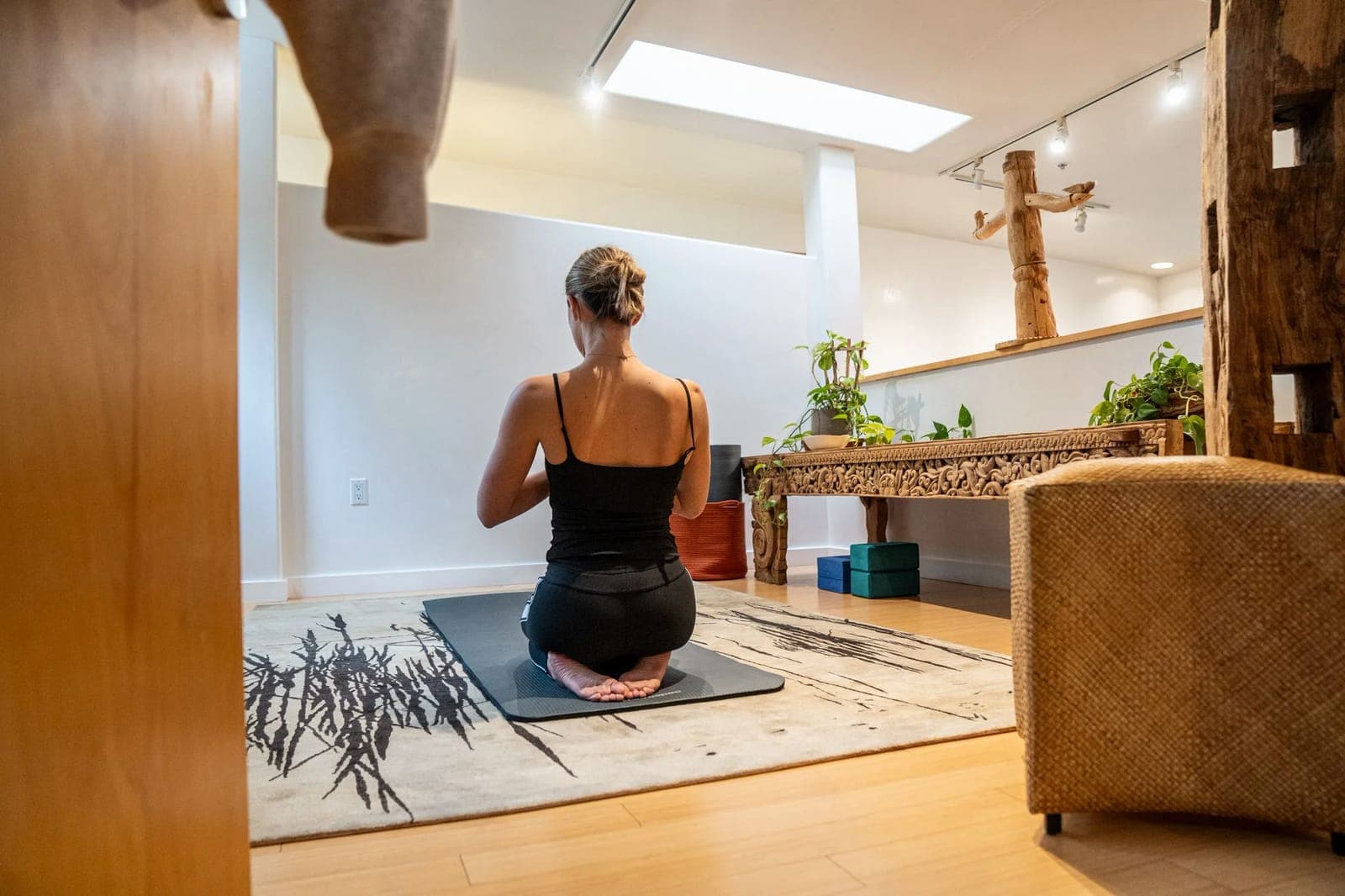 A woman kneeling on a yoga mat in Tumbleweed's sunlit loft, facing a carved wooden console under a skylight