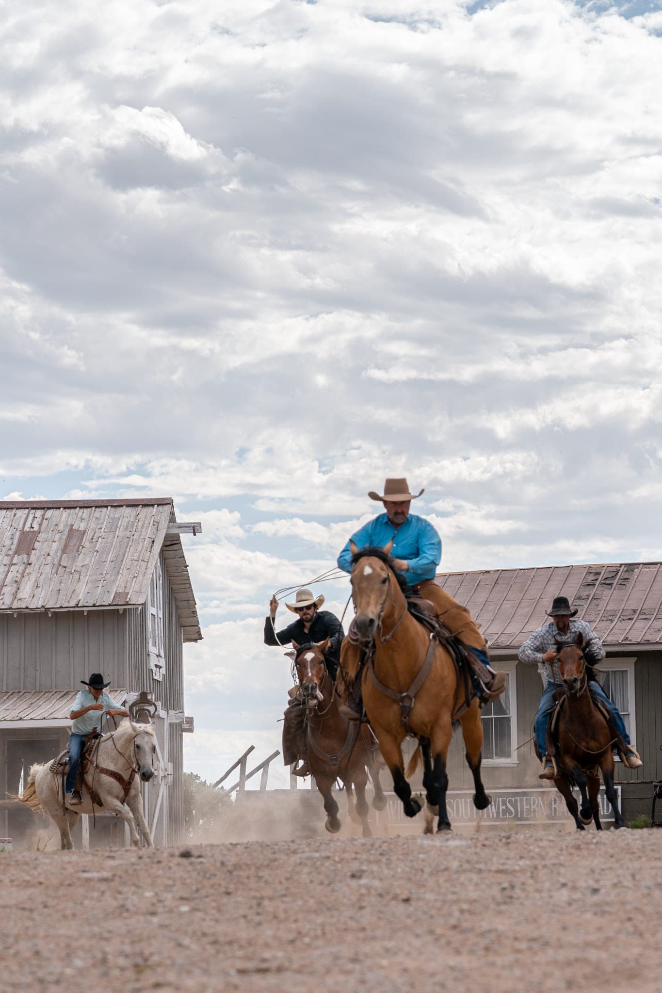 Four riders galloping into a dusty ghost-town street past weathered buildings