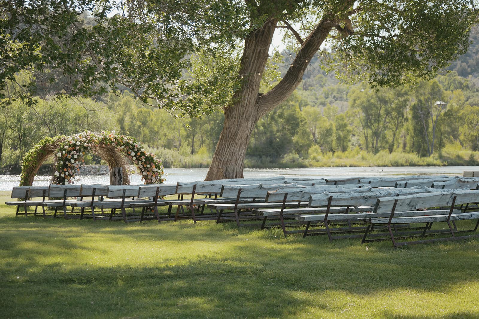 Wooden ceremony benches under a cottonwood facing a floral arch beside the pond at LePlatt’s Pond, Bayfield, Colorado