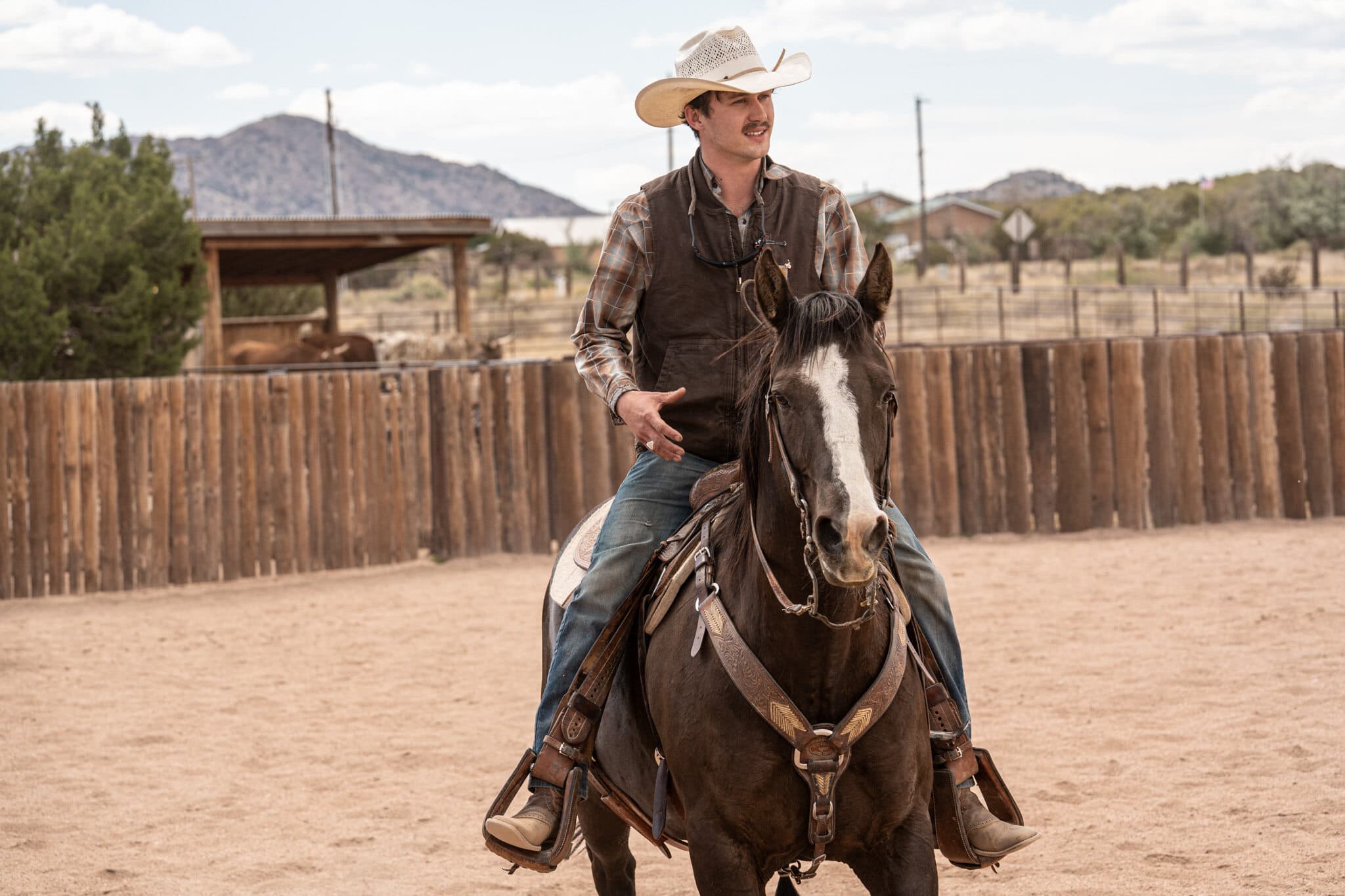 A young cowboy in a white straw hat sitting a dark bay horse in the arena at the 6666 Ranch, mountains behind