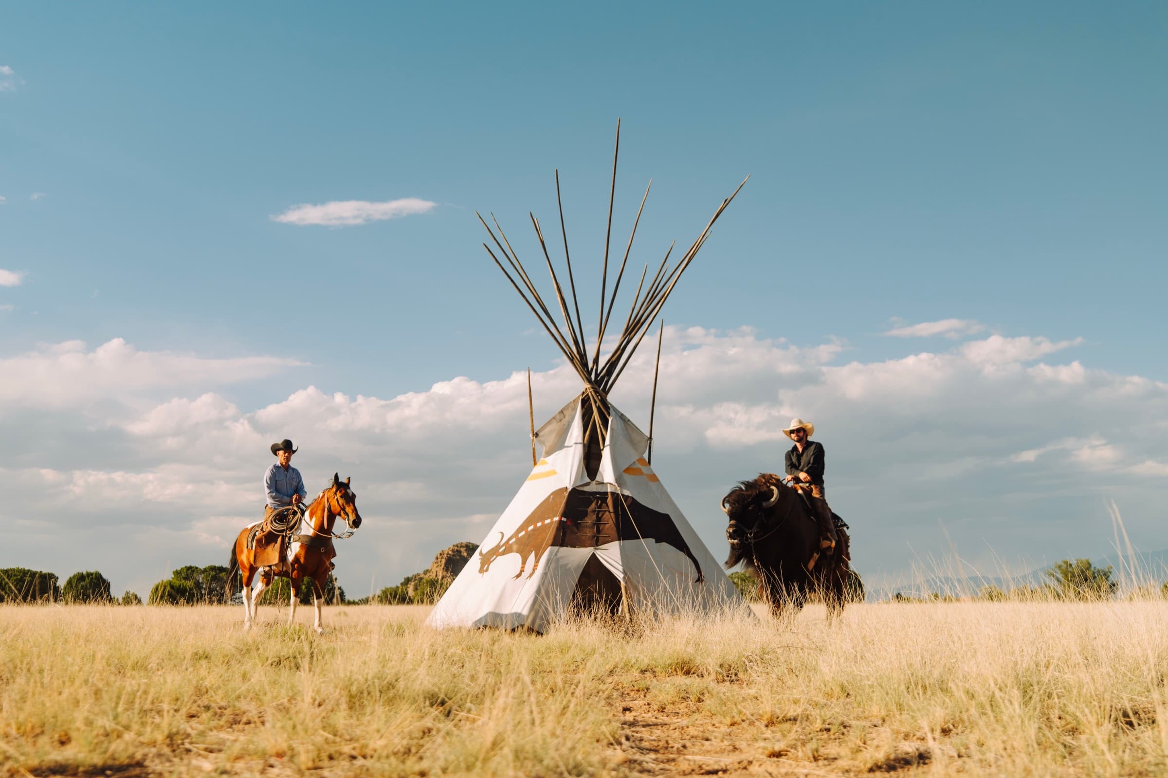 A horse rider and Wyatt Mortenson on Clyde the buffalo flanking a painted tipi in a golden field at the 6666 Ranch