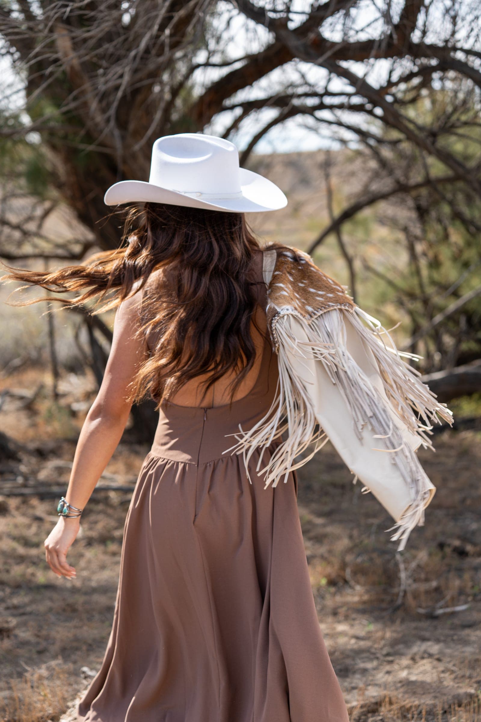 Woman in a white cowboy hat walking away, hair blowing, a fringed hide jacket draped over one shoulder